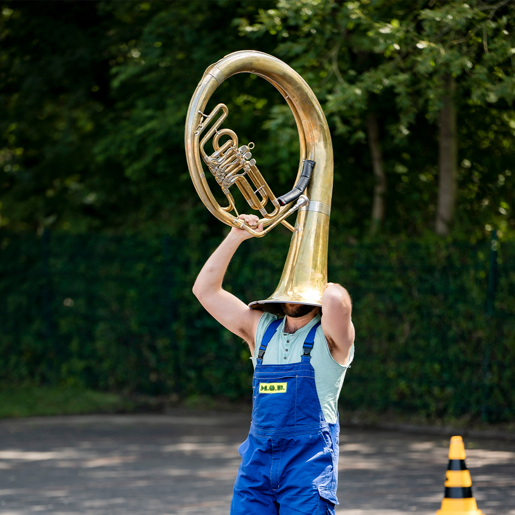 Eine Person im "Blaumann" balanciert eine Tuba auf dem Kopf.