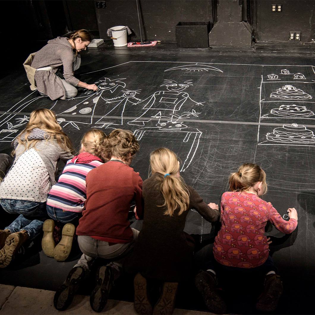5 Kinder und eine Erwachsene Person malen mit Kreise auf schwarzem Bühnenboden. Sie malen Personen in einem Zimmer und Essen.